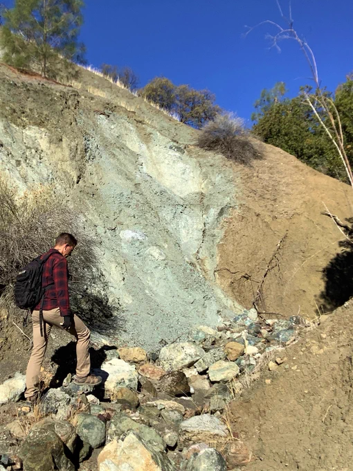 ASU School of Earth and Space Exploration graduate student Joseph Powell in front of the San Andreas fault exposed in a canyon wall. The fault here is marked by white and grey rocks that are highly altered and damaged in the fault zone in up against reddish-brown sediments adjacent to the fault zone. A subtle topographic scarp (the change in slope behind the bush at the top of the fault zone) continues out of view.