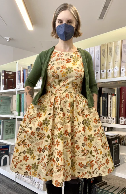 Photo of Jen standing in front of bookshelves, wearing a cream coloured dress with an orange, brown and olive green flower and leaf pattern, a green cardigan and blue mask.