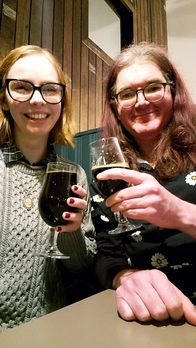 Two ppl in a pub/bar, smiling at the camera and holding beers.