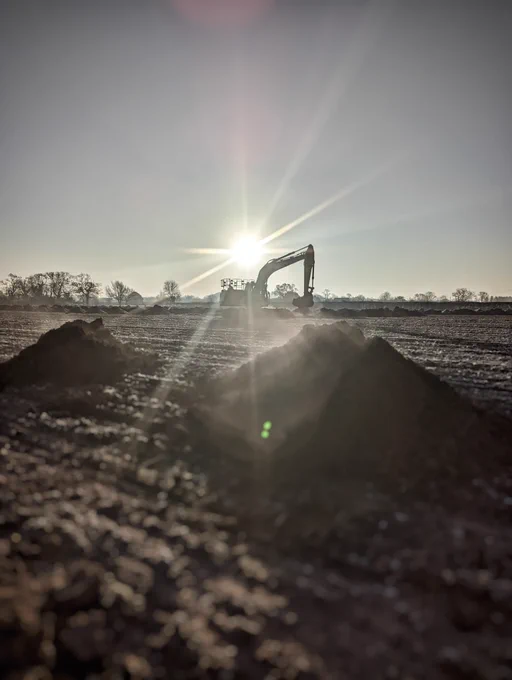 Digger in a frosty ploughed field excavating trenches with the morning sun behind