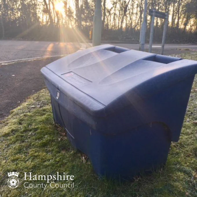 A blue grit bin with a layer of frost on a grass verge by the side of road with the early morning sun shining through the trees in the distance.