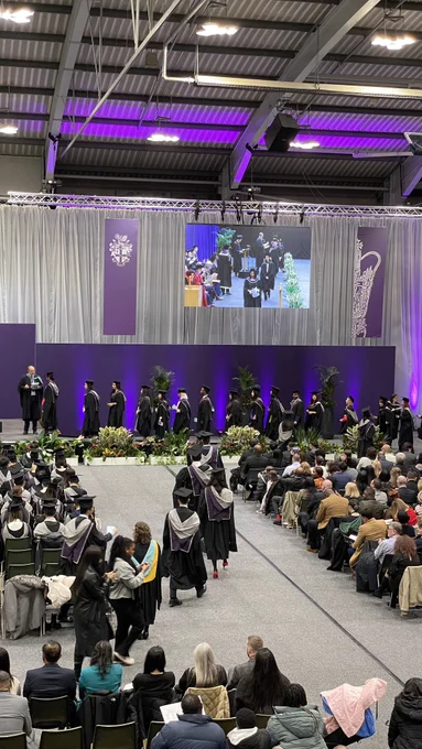 A high view of the graduates walking through the audience towards the stage in a line. They can be seen lined up at the side of the stage. A screen can be seen not he wall, showing a close up of the graduates.