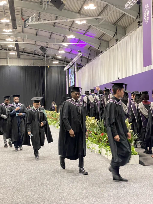 The graduates walking toward the camera, heading towards the stage. Plants line the front of the stage.