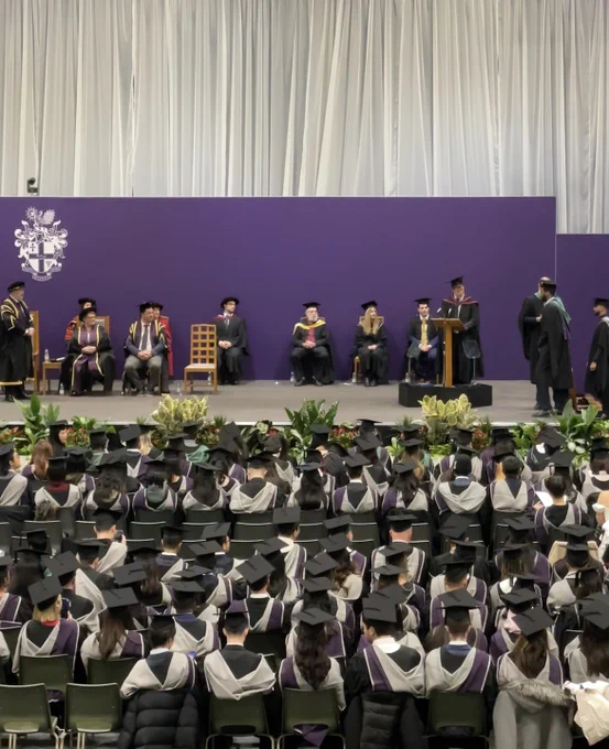A high up shot of the graduates in the audience facing the stage. Other graduates are walking across the stage, with the academics sat at the back of the stage.