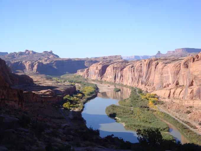 The Colorado River is pictured from above. The river flows through a rocky ravine. Green vegetation grows on either side of the river. The sky is blue and cloudless. 
