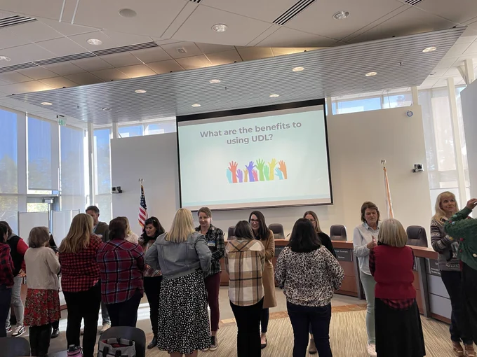 Presentation room with about 20 teachers standing up facing each other in two rows. They are answering question from projector: what are the benefits to using UDL?