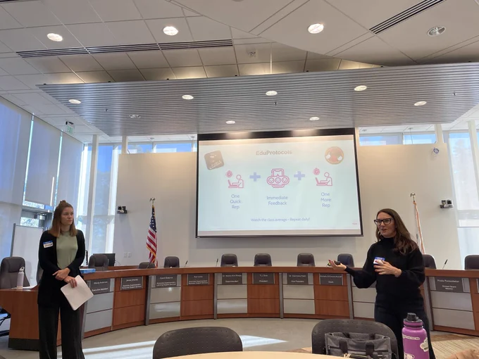 2 white women standing in front of a presentation room on front of a presentation screen