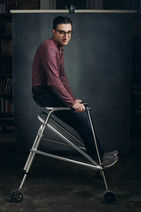 Ryan sits elevated on the back bar of his metallic walker, with the walker itself in profile and Ryan turning his head to give the camera a cheeky grin. He wears a maroon textured Henley, dark jeans, and grey tennis shoes in front of a navy photography backdrop.