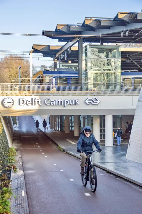 An elderly man in winter attire cycles through an underpass beneath Delft Campus Station. A train waits at the platform above, and the roof is made of black solar panels.
