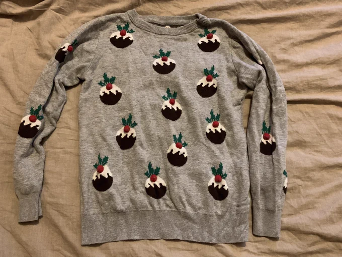A grey jumper with small Christmas puddings dotted over it. The puds are dark brown beneath, white on top, with red pom-poms Holly berries and dark green leaves.
