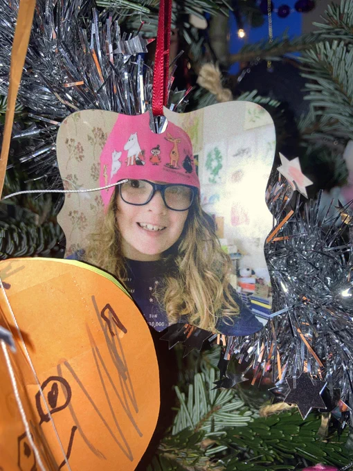 A photo of my daughter Flo wearing a pink beanie hat with Moomins on it, with her long red curly hair showing underneath, her dark rimmed glasses and a dark blue long sleeved top, printed on a metal Christmas tree bauble, next to an orange Christmas tree decoration she has made. Both ornaments are hanging on a real Christmas tree with silver tinsel and white twinkling lights.