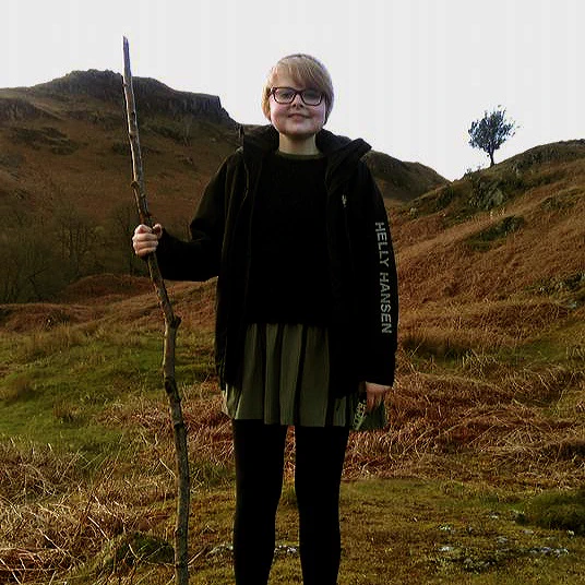 A photograph of Lizzy in a woolly hat and black puffa jacket, smiling triumphantly at the camera whilst holding a large walking stick, in front of a mountain.