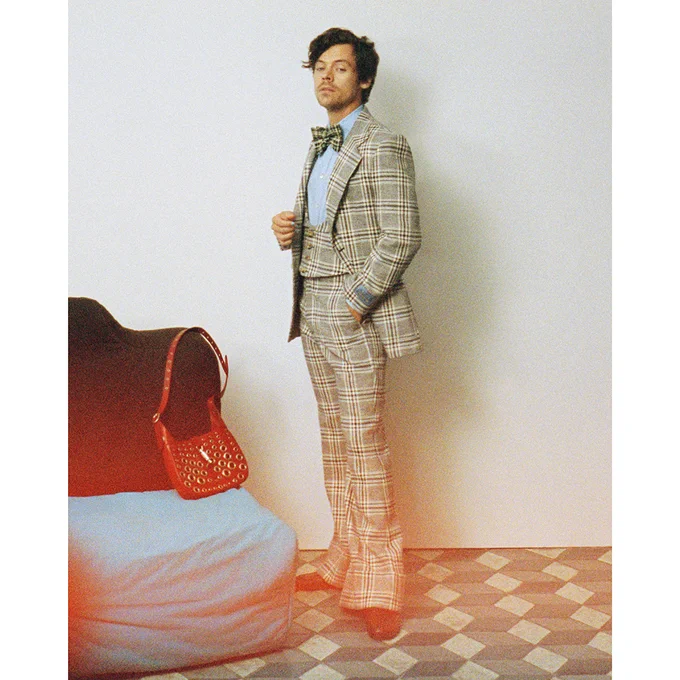 Harry Styles, his wavy brown hair parted on the side, stands with his feet in V, one hand in the pants pockets of his three piece suit, the other held near his low cut vest, in a white room with a cube patterned stone floor. Next to him a large red leather Jackie 1961 bag sits on a couch with its long shoulder strap and all over metal tunnel insert motif. Also wearing a light blue Oxford and large check bowtie, Styles looks down playfully towards the camera.  