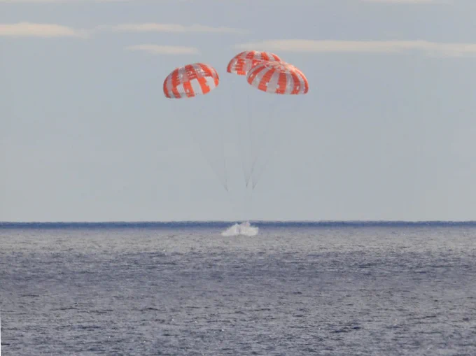 A white splash is visible as three red and white main parachutes lower the Orion crew module into the blue sea off the California coast.