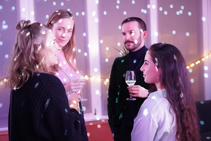 Drei junge Frauen und ein junger Mann stehen in einem hellen Raum; die junge Frau und der junge Mann in der Mitte halten jeweils ein Weinglas in der Hand; im Raum sind viele kleine Lichtpunkte einer Diskokugel zu erkennen; der Blick ist auf die junge Frau links außen gerichtet, die etwas erzählt