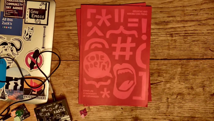 A spanking new code poetry book with cutup graphics on a wooden table, suggestively accompanied by accessories for post-social media lifestyle such as Emacs and synthesizer