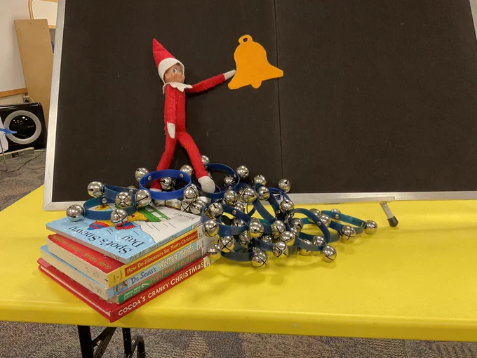 An elf on the shelf stuck to a felt board with a felt bell, a tangle of jingle bells underneath, and a stack of christmas board books