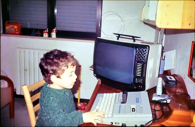 Photo of Nicola as a 4 or 5 year old, sitting in his room in front of a vintage computer: something that looks like a clunky keyboard. Next to him, a catod tube television.