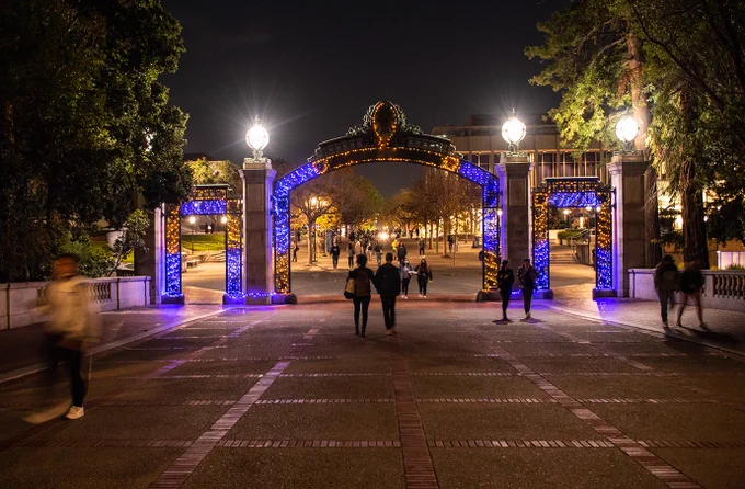 Sather Gate at the University of California, Berkeley. It is night and the gate is decorated in festive blue and gold lights. A few people are walking in the area.