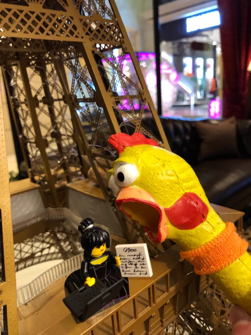 The Lego figure, here seen wearing a dress and a paper, standing on a model Eiffel Tower while the head of a yellow rubber chicken seems to be about to eat her