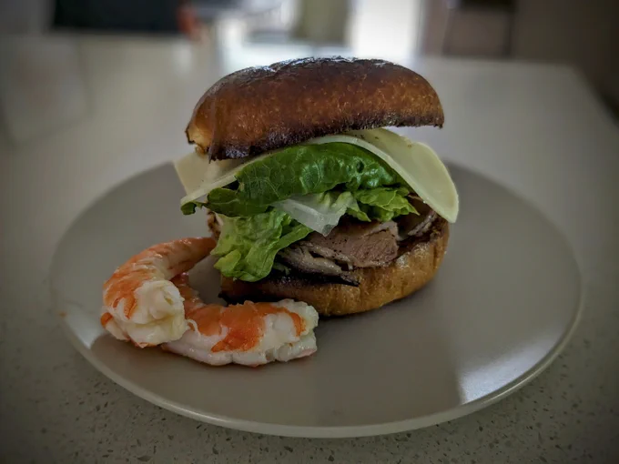 A toasted brioche bun sandwich sits on a small plate. Visible layers are cheese, cos lettuce and fried bacon. Sitting next to the sandwich are two cooked and peeled king prawns.