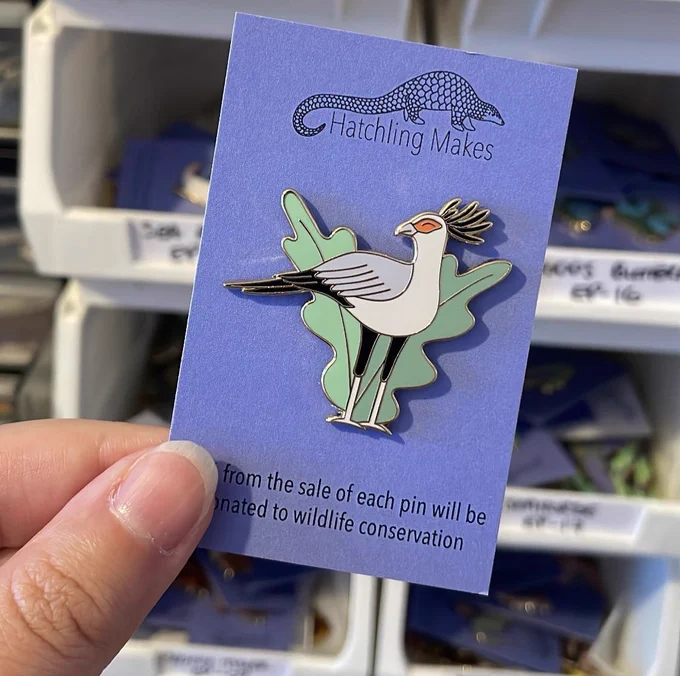 A hand holding an enamel pin of a Secretary bird on a blue backing card
