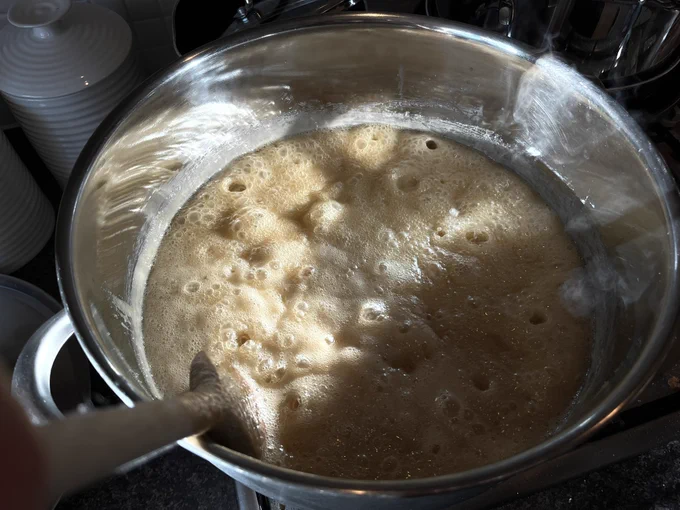 Large metal pan with wooden spoon sitting on the gas ring in winter sunshine. It’s two thirds full with bubbling golden sugar, butter and milk. You can see the bubble craters too