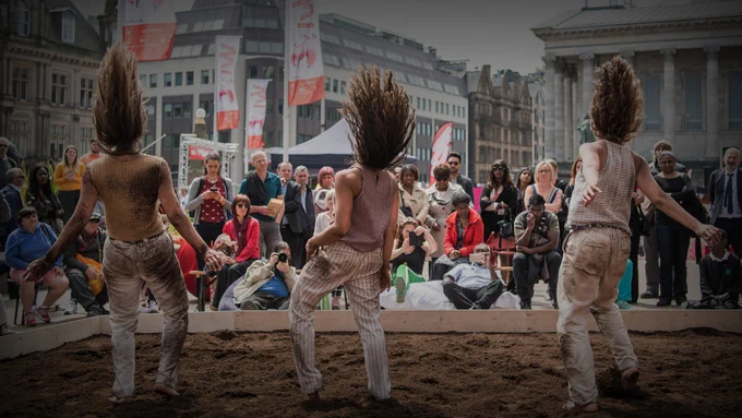 Three people perform outdoors in front of a crowd. The performers have long hair which stretches upwards from their heads.