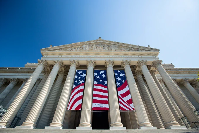 Color photograph of the columns of front portico of the National Archives building on the Constitution Avenue side. Three banners hang between the columns, creating a flag.