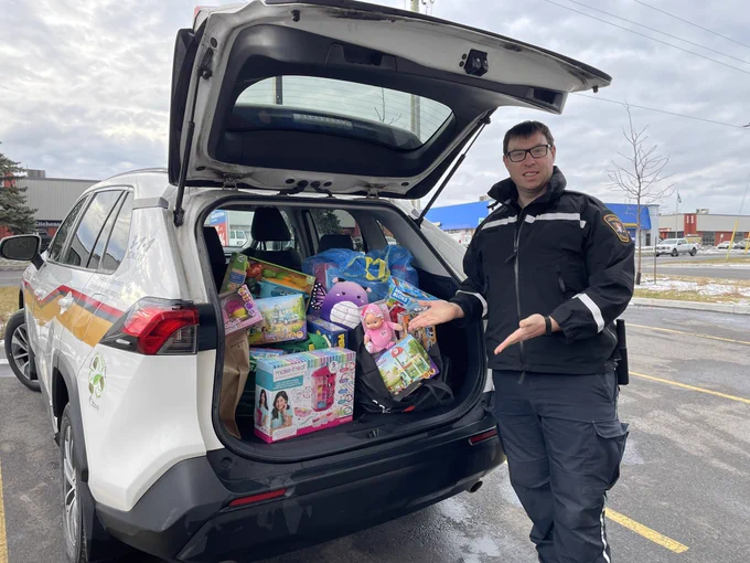 A By-law officer stands next to a vehicle with an open trunk that is filled with toys.