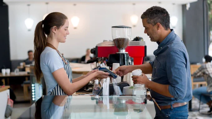 Photo of a customer and employee in a coffee shop. The customer is paying via a contactless card machine