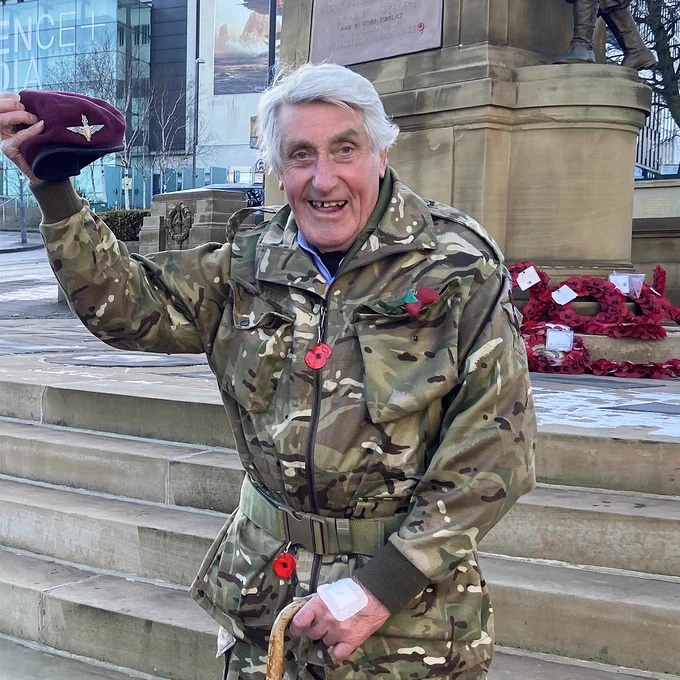 Former paratrooper, Jeffrey Long, stands in front of the Cenotaph in Bradford. He is wearing British Army multi-terrain pattern uniform and holds a walking stick in his left hand. In his right hand, he holds up a maroon beret, adorned with a parachute regiment badge.