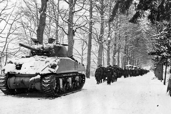 Historic black and white photo of a line of American troops marching behind a tank in snow covered woods.
