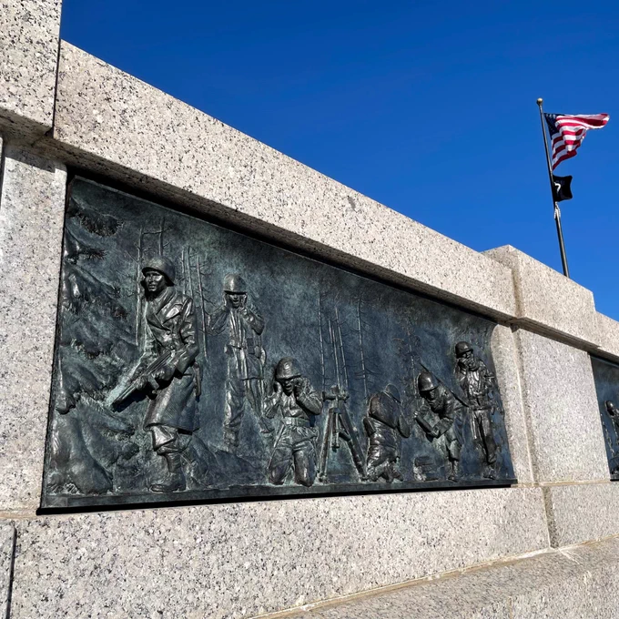 A bronze artistic panel embedded in an outdoor stone memorial wall shows a scene of American soldiers fighting in winter woods during World War II.