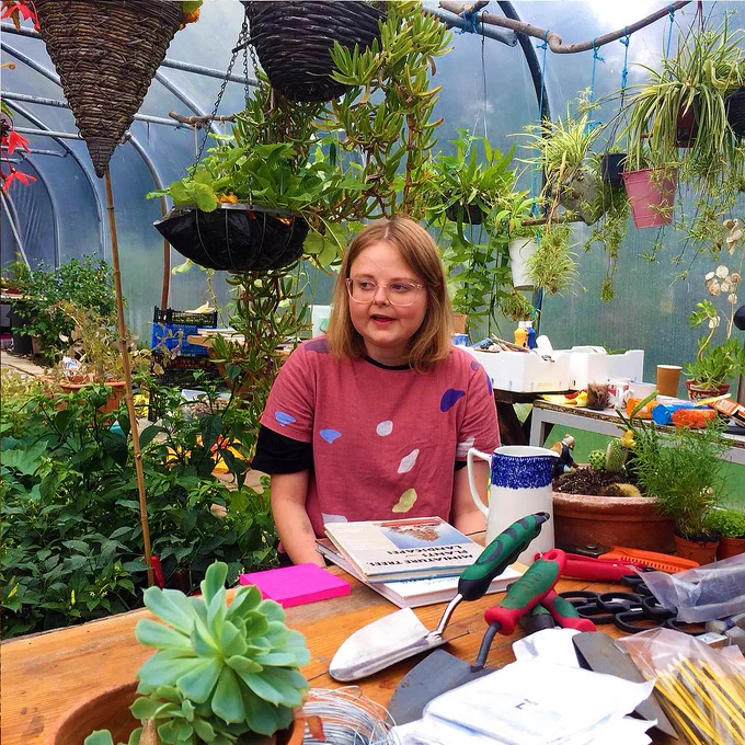 Photo of Lizzy Rose in a large greenhouse surrounded by plants and gardening tools. 