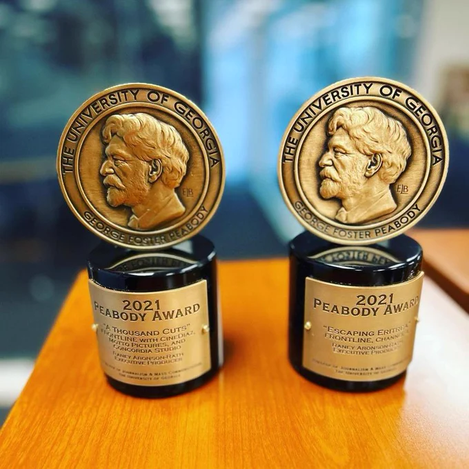 FRONTLINE's two 2021 Peabody Awards for "A Thousand Cuts" and "Escaping Eritrea" sit next to each other on a wooden table. Photo courtesy: FRONTLINE Executive Producer and Editor-in-Chief Raney Aronson-Rath.