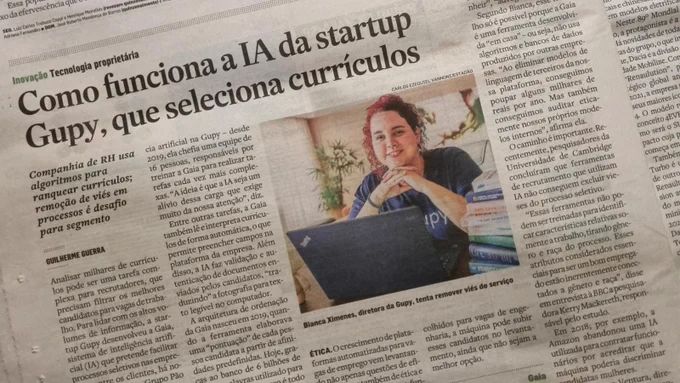 Image of a newspaper article with an image of a woman in front a laptop with headline text that says "Como funciona a IA da startup Gupy, que seleciona curriculos"