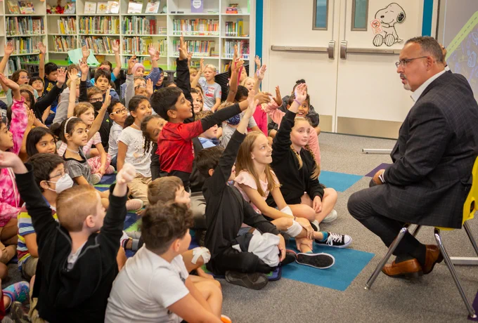 Secretary Cardona speaks with students as they sit around him in a classroom raising their hands
