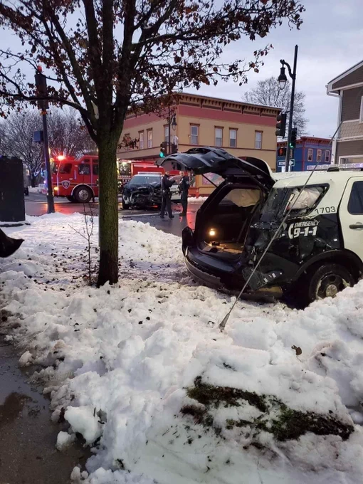 A photo showing two heavily damaged cop cars at the corner of North Street and North Champlain. One is on the curb, has heavy rear end damage, and the airbags are deployed; the other has heavy front-end damage. There is a fire truck in the background and two cops are standing in the middle of the road. There is snow on the ground.