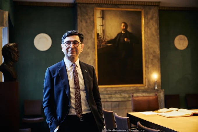 Ardem Patapoutian standing in the Nobel Foundation room with a portrait of Alfred Nobel in the background. He is smiling and wearing a suit.