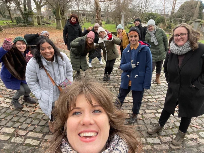 A selfie with the face of a woman with brown hair in her 30s at the front with a group of 12 woman standing behind her smiling at the camera. You can see trees and tomb stones in the background. They are wearing coats, hats and scarfs. There are standing on cobble stones. 