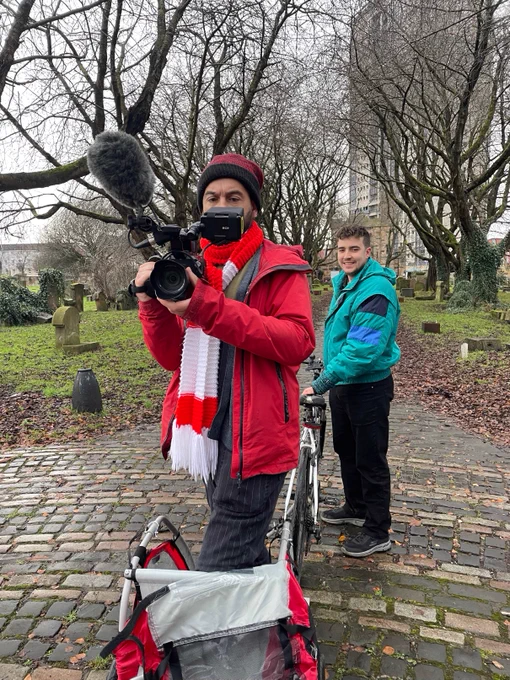 There are two men in the picture. One is at the front, wearing a read coat, hat and red and white scarf. He has a big video camera in his hand which is pointing towards us. He is standing in a small bike trailer. Behind him is a young man in a green coat and black jeans holding a bike. The bike and the trailer are attached. He is standing side ways and smiling at the camera. There are tomb stones and old, trees in the background with no leaves. They are standing on cobble stones. 