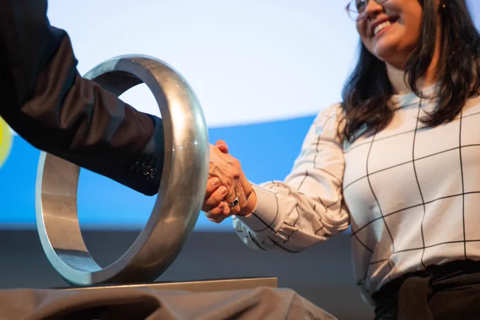 A smiling woman in a white top shakes hands with someone as their hands meet within the oversized ring symbolizing the Order of the Engineer. She is a graduating engineering student.

