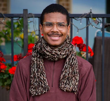 Photo of Aqshems Nichols smiling, wearing glasses, a brown woven scarf, and maroon pullover.