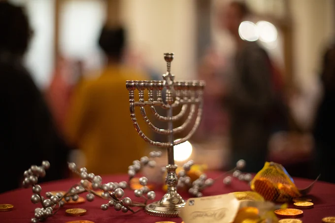 A small menorah is placed on a table with red cloth, winter holiday décor and chocolate gold coins; in the background and out of focus are several people attending a 2018 menorah lighting event