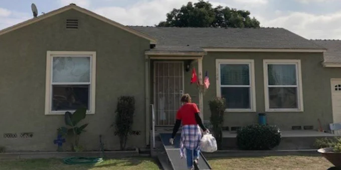 Woman carrying groceries walks to front door of a one-story home.