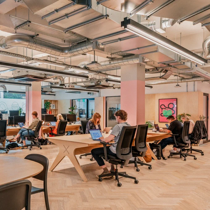 A shot of our new studio. It’s an open, bright space with two central pink pillars. Either side of the pillars are our desks, with Humans sitting at them working. There’s art on the walls. 
