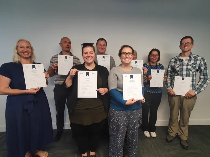 Eight men and women standing inside a room with their backs to a white wall. Each holding up a certificate. Each person is smiling.
