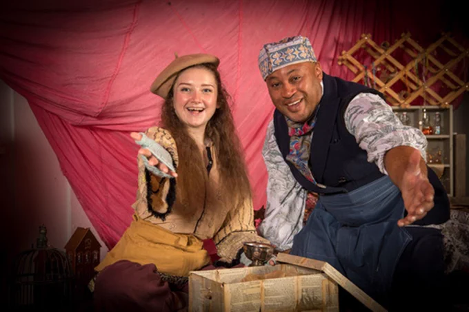 Two friendly actors from the show in costume surrounded by props face the camera with hands outstretched welcoming you to discover more