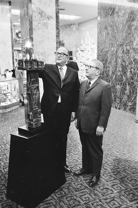 A black & white photo graph of 2 white men standing next to a large menorah on a pedestal. Both of them are wearing suits and glasses. One of them reaches up toward an illuminated light bulb. They are in a lobby of a building with carpeted floors and smooth-stone covered walls. On the left are glass counter & in the background is a Christmas tree.